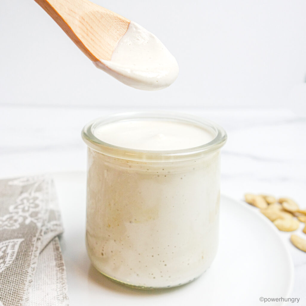 a wooden spoon dipped into a clear glass jar full of cashew cream